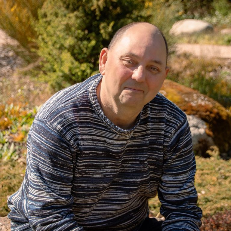 Christian Schwabe Mann mit gestreiftem Pullover sitzt im Freien, umgeben von Pflanzen und Felsen.
